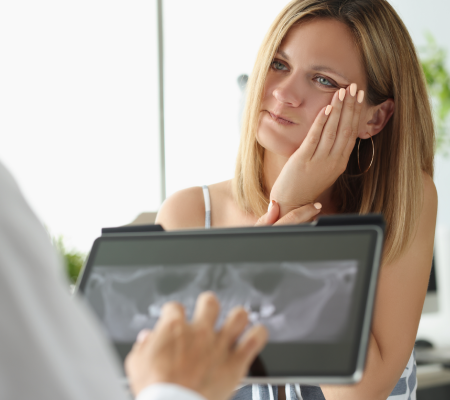 Woman holding her cheek in pain while a dentist reviews her X-ray on a tablet